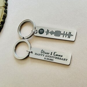 Op de afbeelding: Twee zilveren sleutelhangers. E&eacute;n sleutelhanger heeft een Spotify-code. De andere sleutelhanger heeft de tekst "Oliver & Emma HAPPY ANNIVERSARY 21.08.2021". Beide sleutelhangers hebben een zilveren sleutelring.