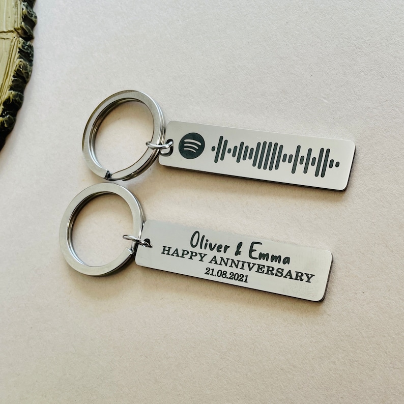 Op de afbeelding: Twee zilveren sleutelhangers. E&eacute;n sleutelhanger heeft een Spotify-code. De andere sleutelhanger heeft de tekst "Oliver & Emma HAPPY ANNIVERSARY 21.08.2021". Beide sleutelhangers hebben een zilveren sleutelring.