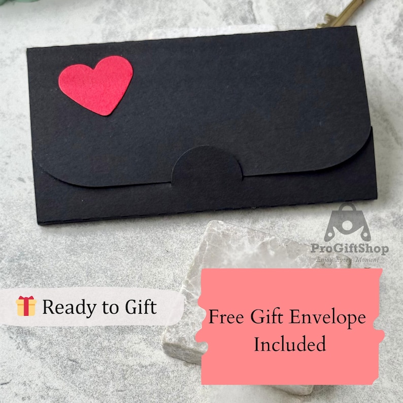 Op de afbeelding: Een zwarte cadeau-envelop met een rode hartvormige accent. De rechthoekige envelop heeft een gebogen flap en een cirkelvormige sluiting. De tekst "Ready to Gift" is zichtbaar.