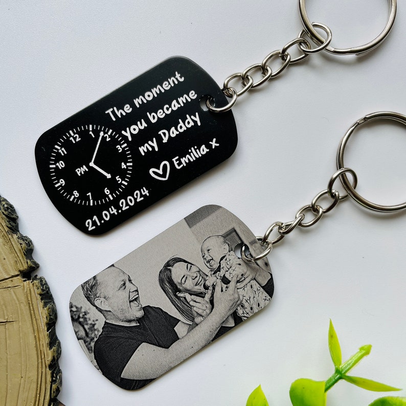 Puede incluir: Dos llaveros rectangulares negros con cadenas plateadas. Un llavero tiene un dise&ntilde;o de reloj y el texto "The moment you became my Daddy Emilia x 21.04.2024". El otro llavero tiene una foto en blanco y negro de una familia.
