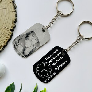 Puede incluir: Dos llaveros personalizados. Uno presenta una foto en blanco y negro de un beb&eacute;. El otro es negro con texto blanco: "The moment you became my Daddy" con un dise&ntilde;o de reloj, la fecha 21.04.2024 y el nombre "Emilia x".