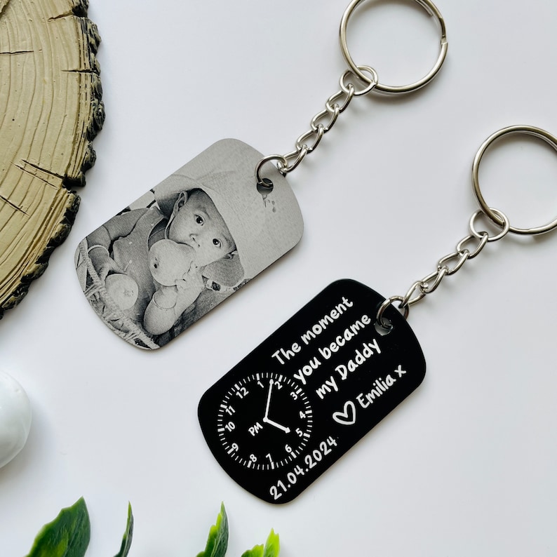 Puede incluir: Dos llaveros personalizados. Uno presenta una foto en blanco y negro de un beb&eacute;. El otro es negro con texto blanco: "The moment you became my Daddy" con un dise&ntilde;o de reloj, la fecha 21.04.2024 y el nombre "Emilia x".