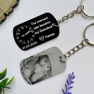 Puede incluir: Dos llaveros personalizados. Un llavero negro presenta un dise&ntilde;o de reloj y el texto "The moment you became my Grandma" con una fecha y un nombre. El otro llavero muestra una foto en blanco y negro de un beb&eacute;.