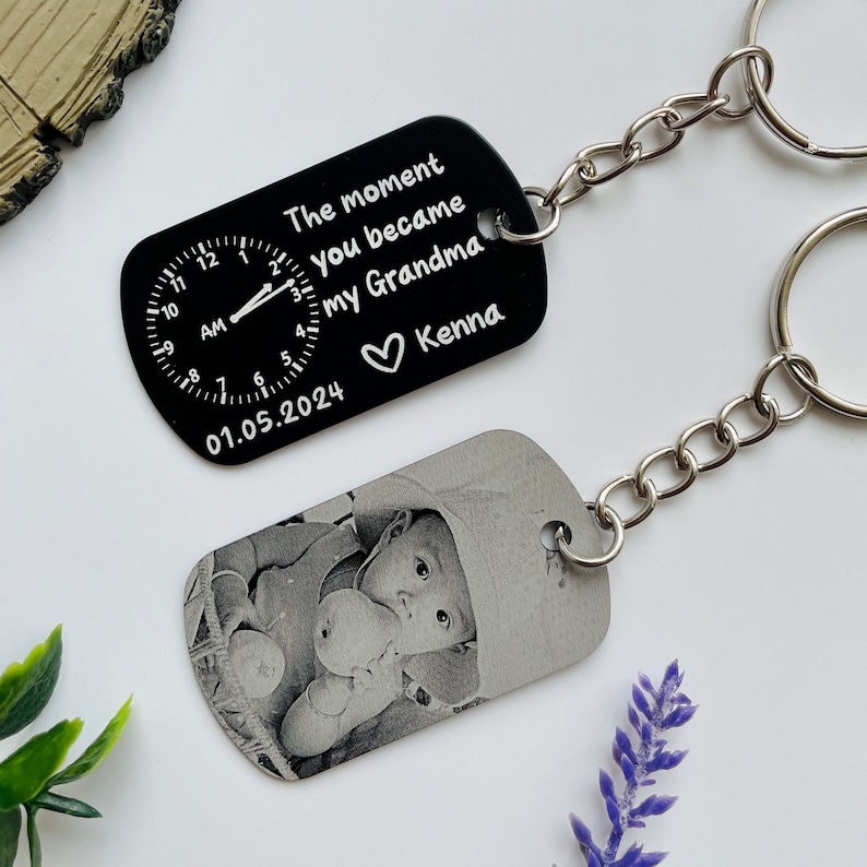 Puede incluir: Dos llaveros personalizados. Un llavero negro presenta un dise&ntilde;o de reloj y el texto "The moment you became my Grandma" con una fecha y un nombre. El otro llavero muestra una foto en blanco y negro de un beb&eacute;.