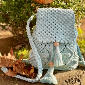 May include: A pale blue macramé backpack with a flap closure. The handmade bag features a decorative floral pattern on the front and a tassel with wooden beads. The shoulder straps are braided.