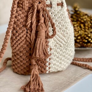 May include: A macrame bucket bag in brown and cream. The bag has a drawstring closure and a long strap. Decorative tassels hang from the front. The bag is made of a thick, woven material.