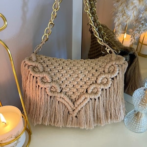May include: A beige macrame shoulder bag with a gold chain strap. The bag has a woven design and a fringed bottom. The chain strap is gold. The bag is on a white surface.