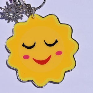 May include: A yellow sun-shaped keychain with a smiling face, closed eyes, and pink cheeks. The keychain has a silver border and is attached to a silver keyring with a small sun charm.