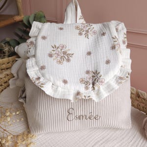 May include: A beige corduroy backpack featuring a white muslin flap with a ruffled edge and a brown floral print. The name Esm&eacute;e is embroidered in brown thread on the front of the bag. The backpack has a top handle and a soft, textured fabric.