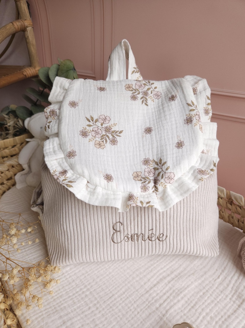 May include: A beige corduroy backpack featuring a white muslin flap with a ruffled edge and a brown floral print. The name Esm&eacute;e is embroidered in brown thread on the front of the bag. The backpack has a top handle and a soft, textured fabric.