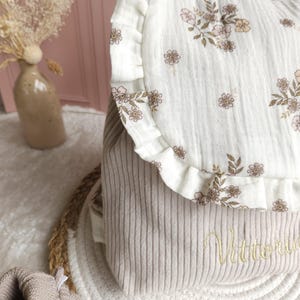 May include: A beige corduroy backpack featuring a white muslin flap with a delicate brown and pink floral print and ruffled edges. The lower section of the backpack is embroidered with the name Vittoria in a cursive gold thread script.