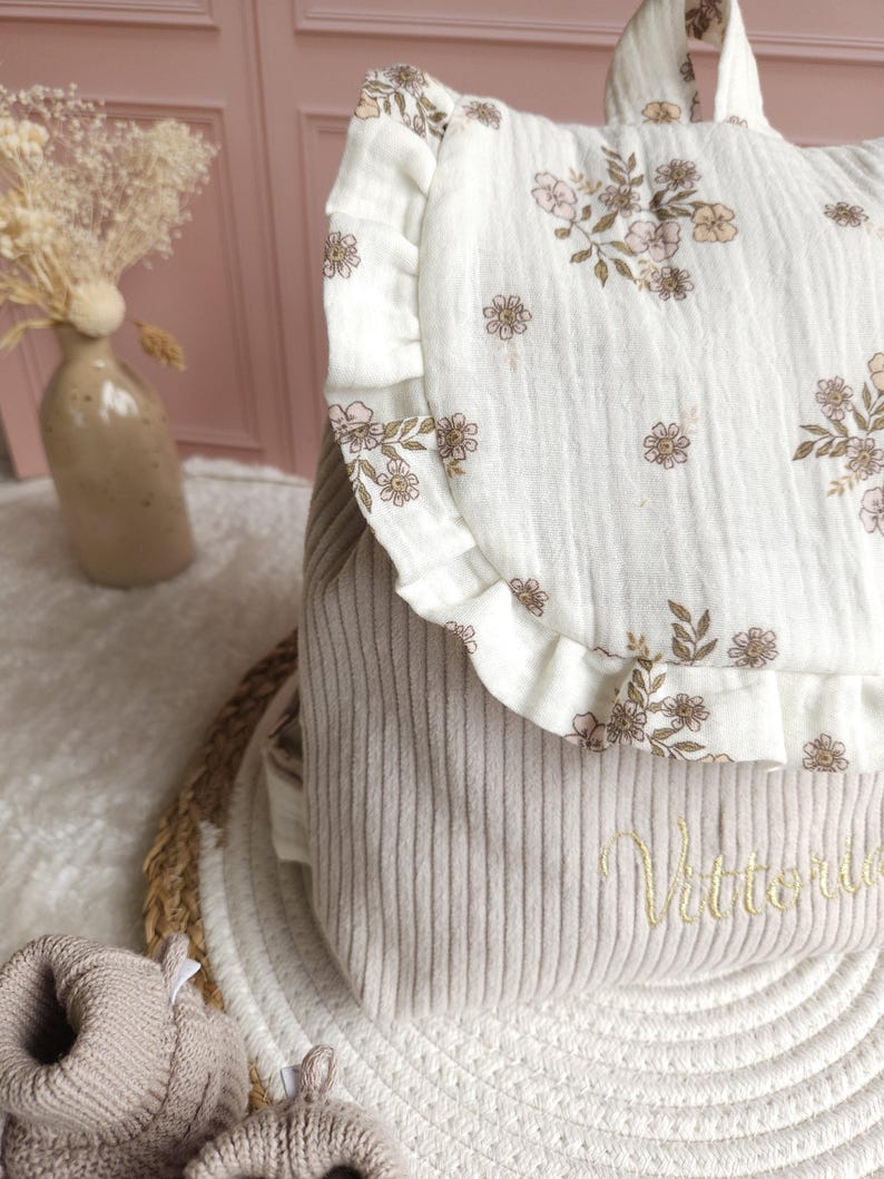 May include: A beige corduroy backpack featuring a white muslin flap with a delicate brown and pink floral print and ruffled edges. The lower section of the backpack is embroidered with the name Vittoria in a cursive gold thread script.