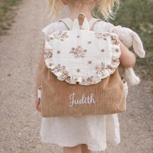 May include: A tan corduroy backpack featuring a cream-colored flap with a delicate brown floral print and a ruffled edge. The name Judith is embroidered in white cursive script on the front pocket. The backpack has matching tan shoulder straps.
