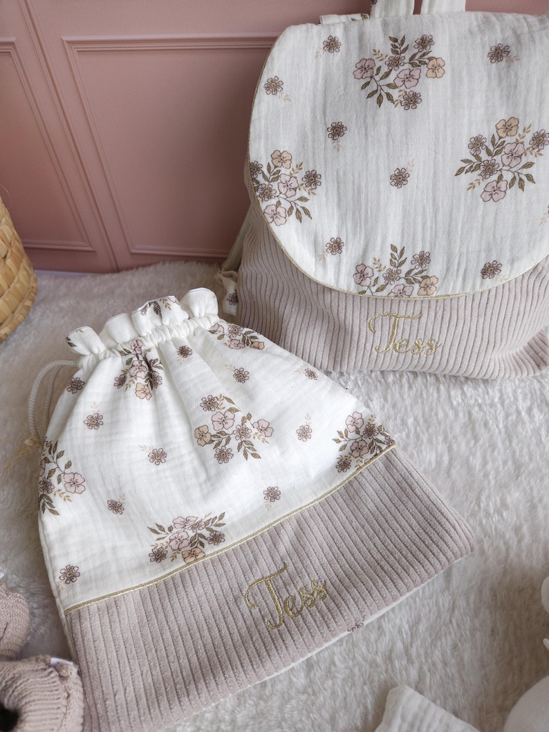 May include: A beige corduroy backpack and a matching drawstring bag, both featuring a white muslin fabric top with a delicate brown and pink floral print. Each item is embroidered with the name Tess in gold thread on the corduroy section.