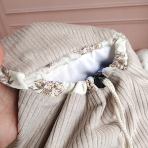 May include: A beige corduroy drawstring bag featuring a white interior lining with a delicate floral pattern in shades of pink and green. The bag includes a white rope drawstring with a black plastic toggle and a silver-toned magnetic snap closure.