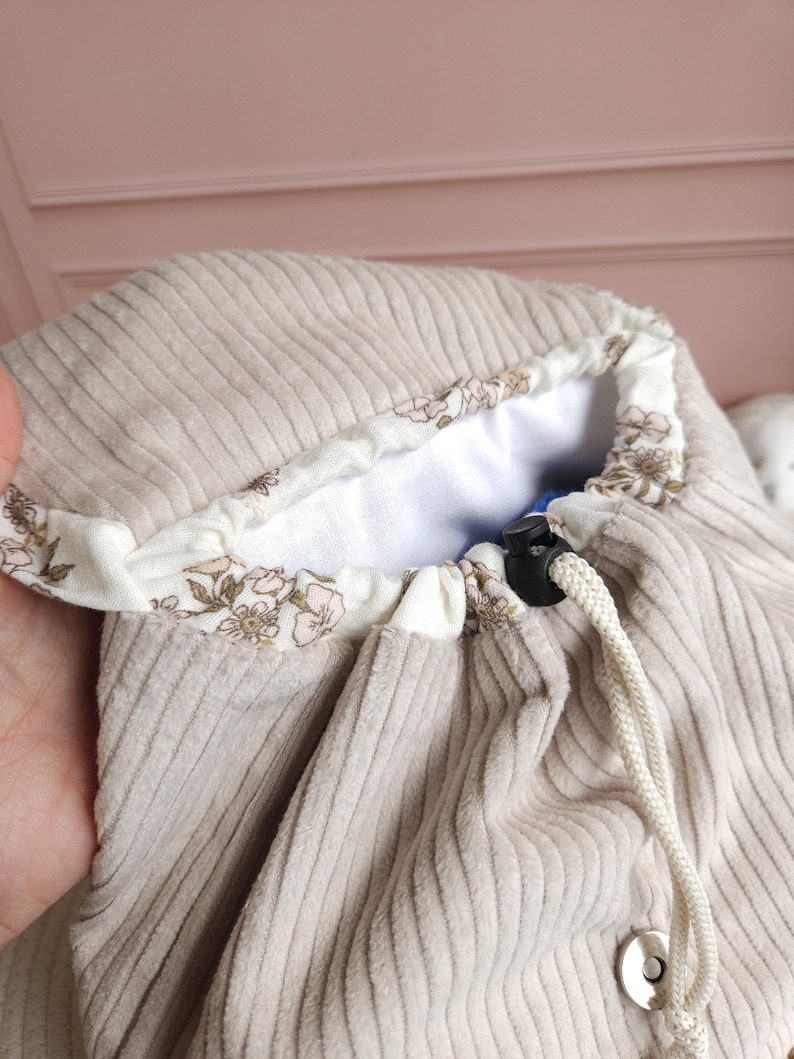 May include: A beige corduroy drawstring bag featuring a white interior lining with a delicate floral pattern in shades of pink and green. The bag includes a white rope drawstring with a black plastic toggle and a silver-toned magnetic snap closure.