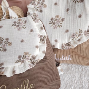 May include: Two corduroy backpacks featuring a white muslin flap with a floral print and ruffled edges. The dark brown backpack is embroidered with the name Camille in gold, while the tan backpack features the name Judith in white embroidery.