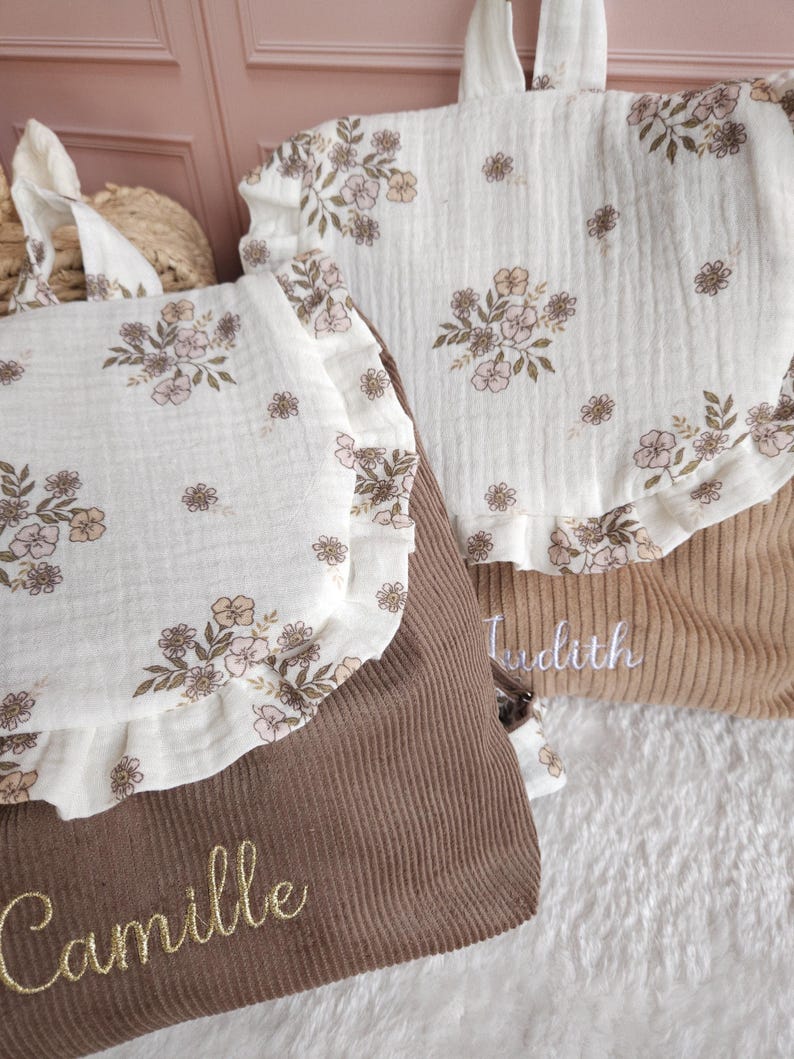 May include: Two corduroy backpacks featuring a white muslin flap with a floral print and ruffled edges. The dark brown backpack is embroidered with the name Camille in gold, while the tan backpack features the name Judith in white embroidery.