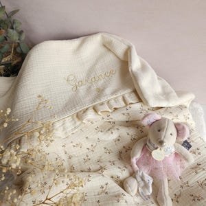 Pode incluir: Um cobertor de beb&ecirc; de musselina creme com um padr&atilde;o floral e o nome "Garance" bordado em fio dourado. Um rato de pel&uacute;cia creme vestindo um tutu rosa est&aacute; sentado no cobertor.