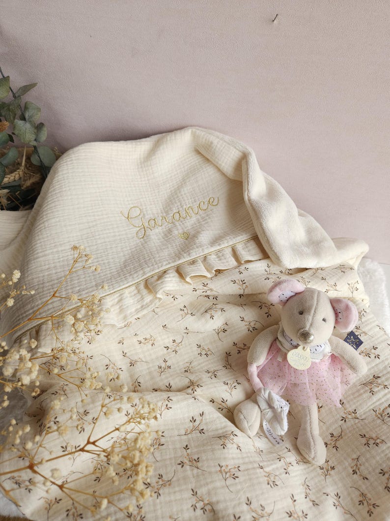 Pode incluir: Um cobertor de beb&ecirc; de musselina creme com um padr&atilde;o floral e o nome "Garance" bordado em fio dourado. Um rato de pel&uacute;cia creme vestindo um tutu rosa est&aacute; sentado no cobertor.