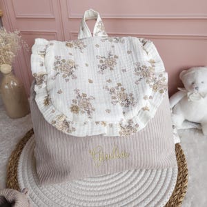 May include: A beige corduroy backpack featuring a white muslin flap with a ruffled edge and a floral print in muted tones. The front of the bag is embroidered with the name Giulia in gold thread, and it includes a top handle for carrying.