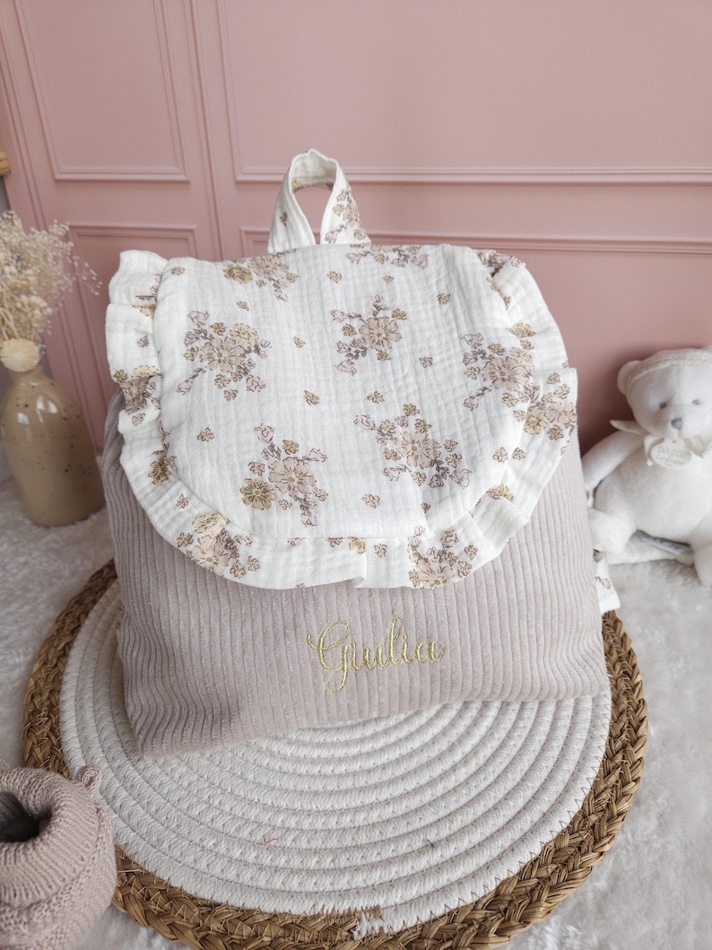May include: A beige corduroy backpack featuring a white muslin flap with a ruffled edge and a floral print in muted tones. The front of the bag is embroidered with the name Giulia in gold thread, and it includes a top handle for carrying.
