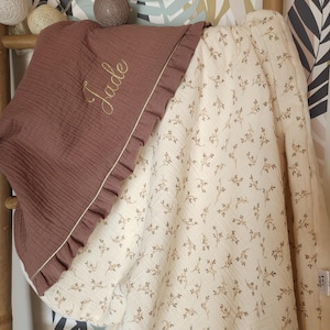 Pode incluir: Um cobertor de muselina marrom para beb&ecirc; com um lado de muselina branco com um pequeno padr&atilde;o floral. O lado marrom tem uma borda com babados e &eacute; bordado com o nome "Jade".