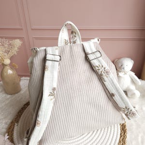 May include: A beige corduroy backpack featuring adjustable shoulder straps with a white fabric overlay printed with a delicate brown floral pattern. The backpack includes a top handle and silver-toned metal hardware for adjusting the strap length.
