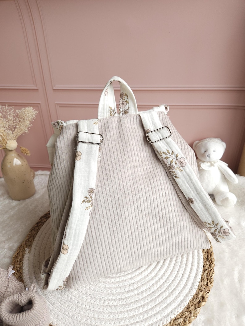 May include: A beige corduroy backpack featuring adjustable shoulder straps with a white fabric overlay printed with a delicate brown floral pattern. The backpack includes a top handle and silver-toned metal hardware for adjusting the strap length.