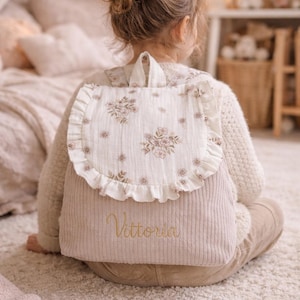 May include: A beige corduroy backpack featuring a white flap with a delicate brown floral print and ruffled edges. The lower section of the bag is made of ribbed fabric and displays the embroidered name Vittoria in a cursive gold script font.