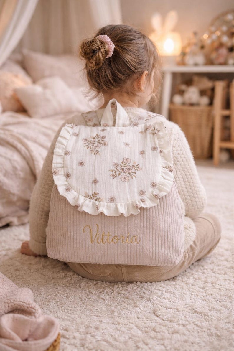 May include: A beige corduroy backpack featuring a white flap with a delicate brown floral print and ruffled edges. The lower section of the bag is made of ribbed fabric and displays the embroidered name Vittoria in a cursive gold script font.