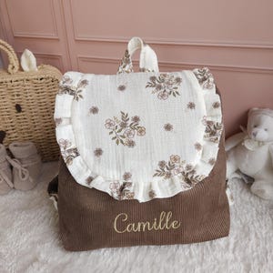 May include: A brown corduroy backpack featuring a cream-colored flap with a delicate floral print and ruffled edges. The name Camille is embroidered in gold thread on the front of the brown corduroy body of the bag, which includes a top handle.