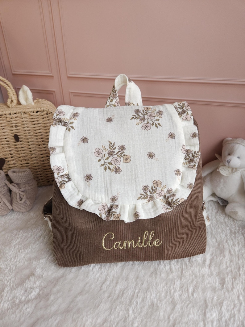May include: A brown corduroy backpack featuring a cream-colored flap with a delicate floral print and ruffled edges. The name Camille is embroidered in gold thread on the front of the brown corduroy body of the bag, which includes a top handle.