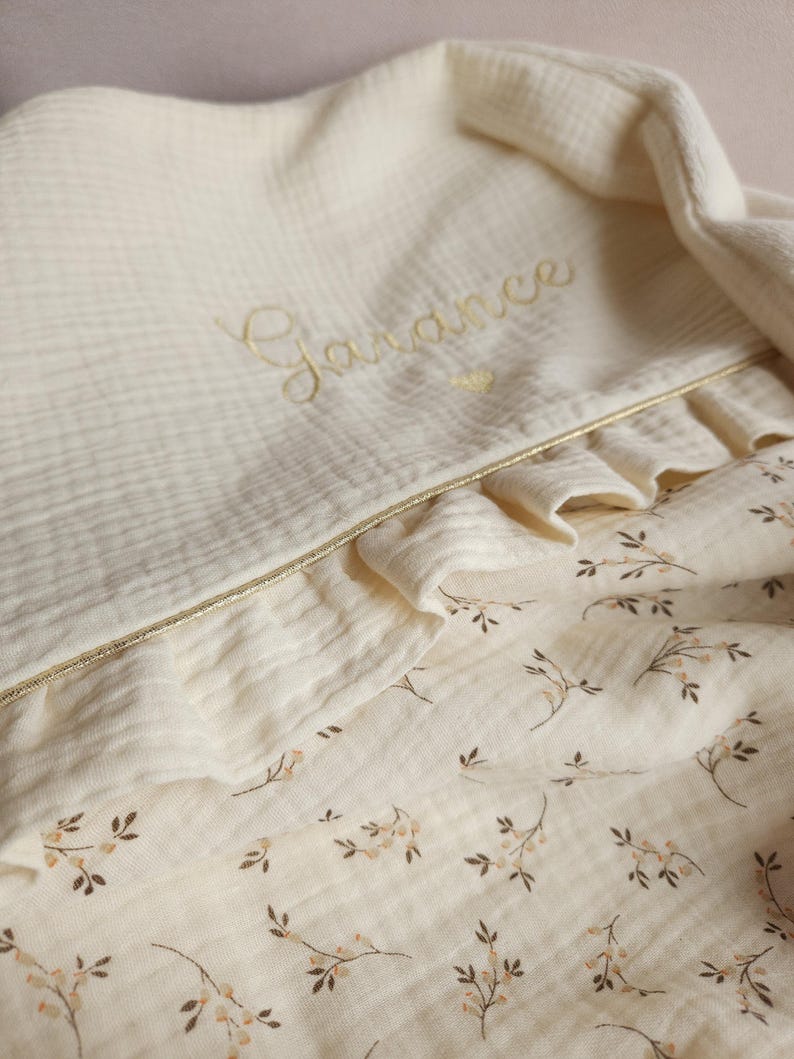 Pode incluir: Um cobertor de muselina branco para beb&eacute; com um delicado padr&atilde;o floral e um nome bordado a ouro "Garance".