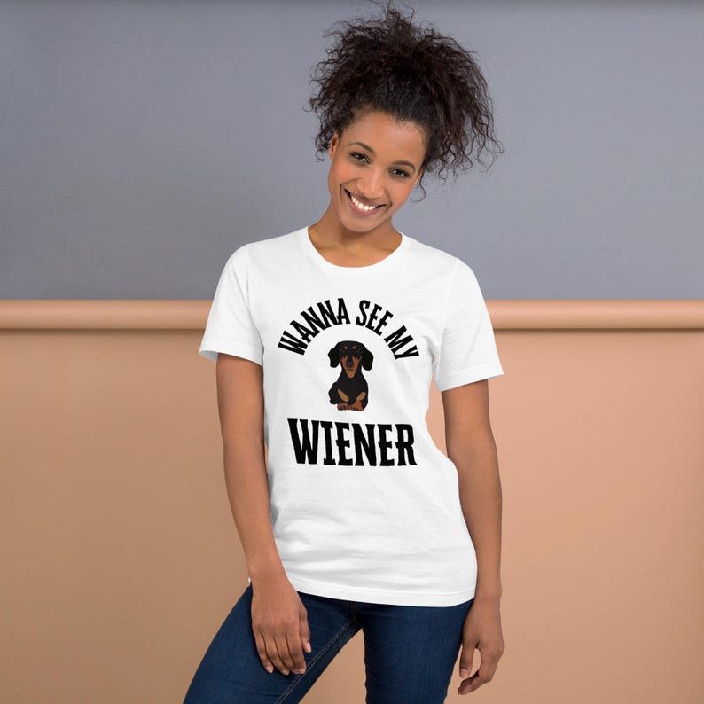 May include: White t-shirt with the text "Wanna See My Wiener" in black, featuring a cartoon dachshund. The dachshund is black and brown. The model is wearing blue jeans.
