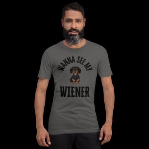May include: A gray t-shirt with the phrase "WANNA SEE MY WIENER" in black block letters. A cartoon dachshund dog is centered between the words. The dog is black and brown with a white chest. The shirt is short-sleeved.