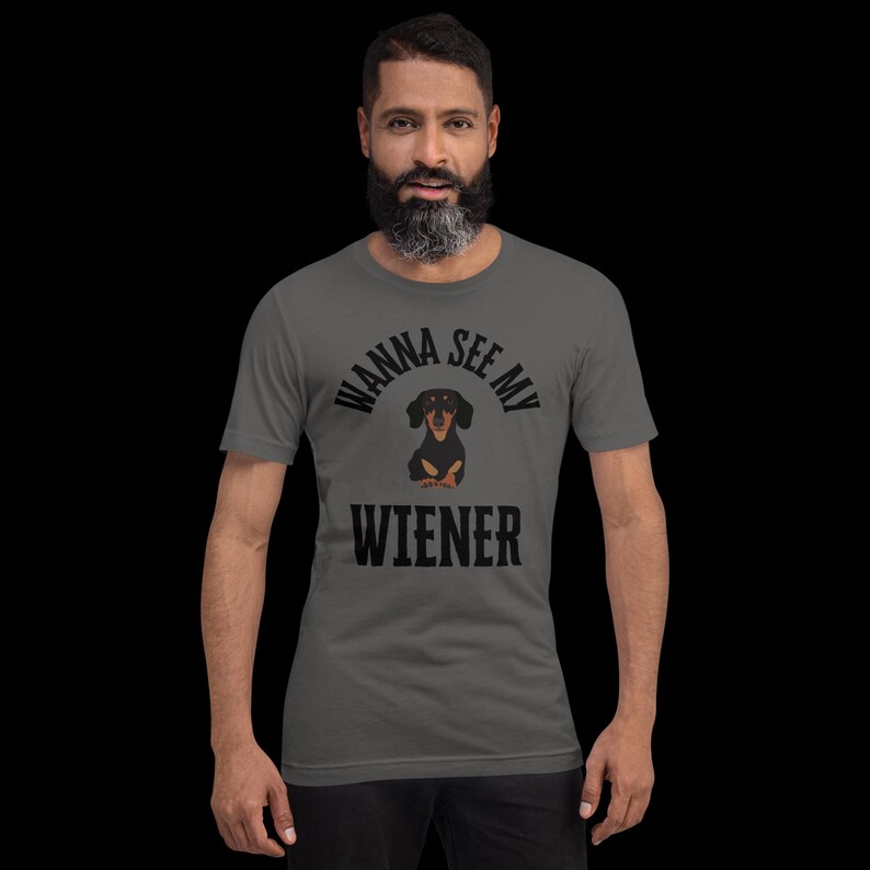 May include: A gray t-shirt with the phrase "WANNA SEE MY WIENER" in black block letters. A cartoon dachshund dog is centered between the words. The dog is black and brown with a white chest. The shirt is short-sleeved.