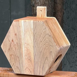 May include: A wooden birdhouse with a geometric design. The birdhouse is made of light-colored wood and has a rectangular top with a small, square perch. A circular opening is visible on the side.