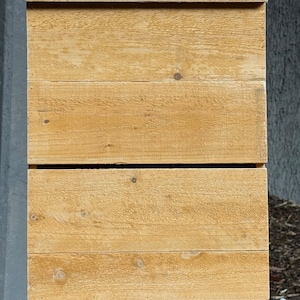 May include: A wooden birdhouse with a rectangular shape and a light brown color. The birdhouse is made of horizontal wooden planks and has a small roof. It is mounted on a tree trunk.