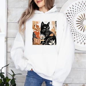 Puede incluir: Sudadera con capucha blanca con una ilustraci&oacute;n de un gato negro rodeado de hojas de oto&ntilde;o y una calabaza. El dise&ntilde;o utiliza tonos negros, naranjas y marrones. La sudadera tiene un bolsillo delantero y una capucha con cord&oacute;n.