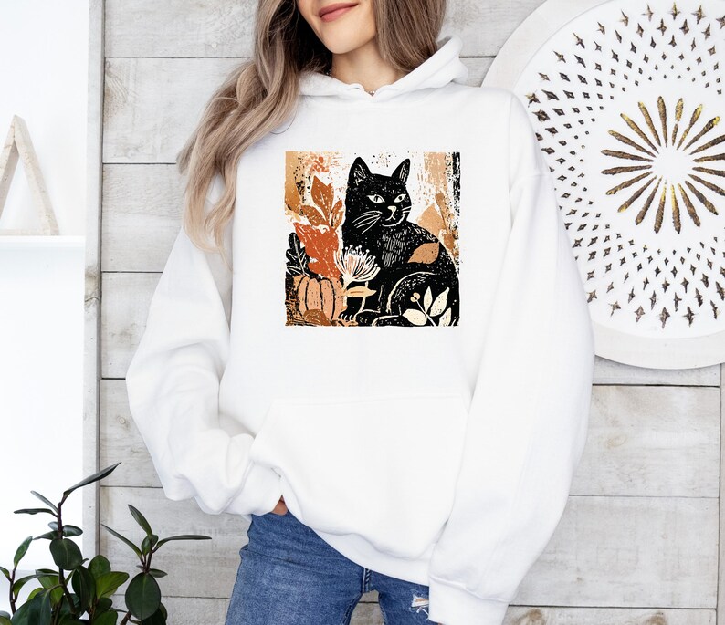 Puede incluir: Sudadera con capucha blanca con una ilustraci&oacute;n de un gato negro rodeado de hojas de oto&ntilde;o y una calabaza. El dise&ntilde;o utiliza tonos negros, naranjas y marrones. La sudadera tiene un bolsillo delantero y una capucha con cord&oacute;n.