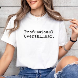 May include: White t-shirt with the text "Professional Overthinker" in black, sans-serif font. The shirt is short-sleeved and appears to be made of a soft material. The person is wearing blue jeans.