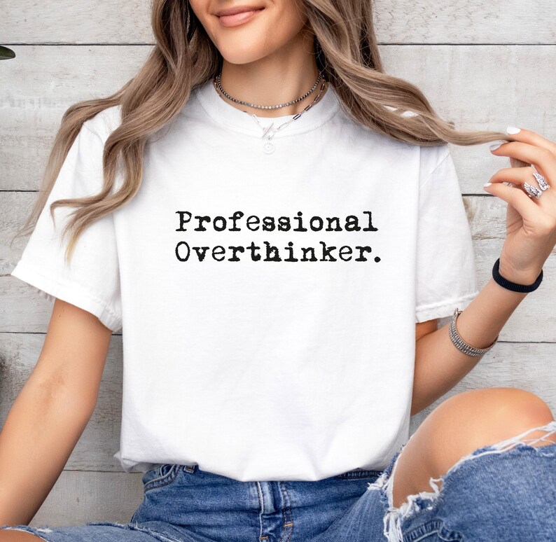 May include: White t-shirt with the text "Professional Overthinker" in black, sans-serif font. The shirt is short-sleeved and appears to be made of a soft material. The person is wearing blue jeans.