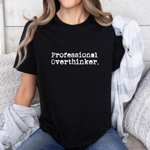 May include: Black t-shirt with the text "Professional Overthinker" in a white, typewriter-style font. The shirt is a crew neck and is being worn by a person. A light-colored plaid shirt is draped over the person's shoulders.