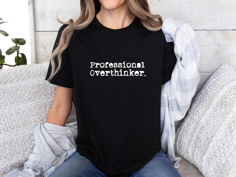 May include: Black t-shirt with the text "Professional Overthinker" in a white, typewriter-style font. The shirt is a crew neck and is being worn by a person. A light-colored plaid shirt is draped over the person's shoulders.