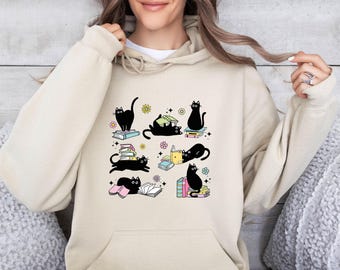 Puede incluir: Una sudadera con capucha de color beige que presenta un estampado gráfico de varios gatos negros interactuando con pilas de libros coloridos. Los gatos aparecen en varias posturas, sentados, tumbados y sobre libros, con pequeños iconos de flores y estrellas.