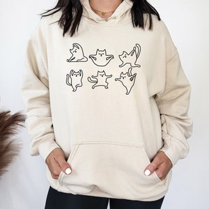 May include: A cream-colored hoodie with a black outline of six cats in different yoga poses. The hoodie has a large front pocket.