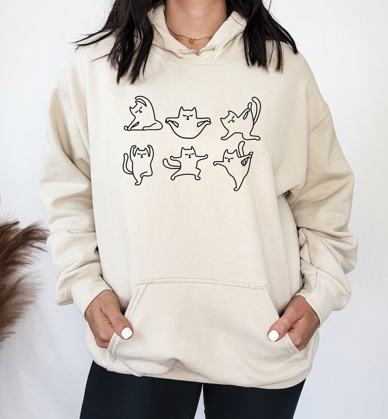 May include: A cream-colored hoodie with a black outline of six cats in different yoga poses. The hoodie has a large front pocket.