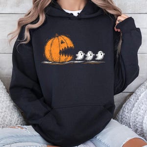 Może przedstawiać: Czarna bluza z kapturem z motywem Halloween. Grafika przedstawia pomarańczową dynię z dużymi, otwartymi ustami, która wydaje się pożerać trzy małe białe duszki. Ilustracja ma styl ręcznie rysowany.