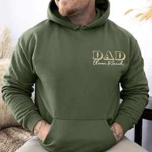 May include: Olive green hoodie with a front pocket and a hood. The text "DAD" is printed in a tan color, with "Oliver & Sarah" below it. The hoodie is made of a soft material.